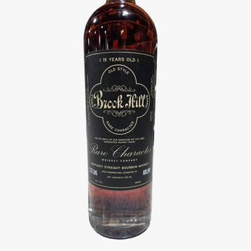 A bottle of Brock Mill Pure Character 15 Years Old bourbon whiskey standing on a speckled countertop with a blurred liquor store shelf in the background.