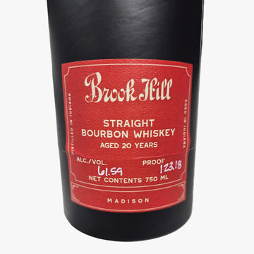 A close-up of a Buck Feill Straight Bourbon Whiskey bottle, showing the black glass body and a red label with white text, resting on a gray marble surface.