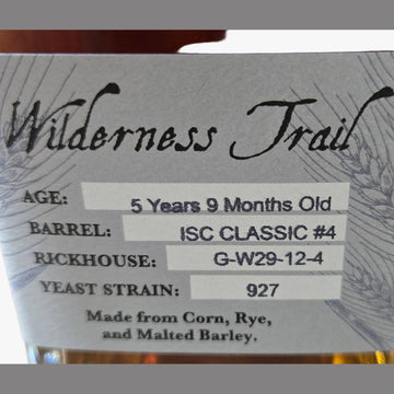 A close-up of a whiskey bottle with amber liquid and a white label reading Wilderness Trail, showing details like age, barrel, and yeast strain.