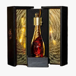 A premium bottle of cognac with a gold decanter cap and red liquid, displayed inside an ornate black and gold gift box on a wooden barrel, with blurred whiskey barrels in the background.