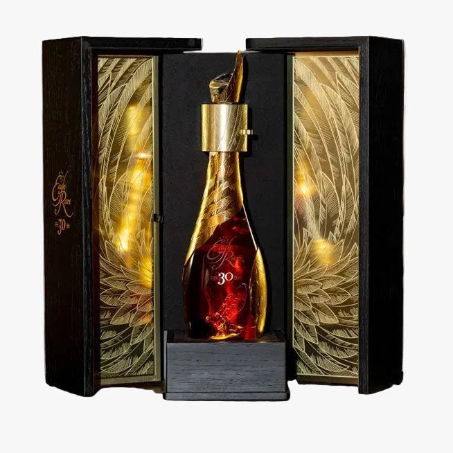 A premium bottle of cognac with a gold decanter cap and red liquid, displayed inside an ornate black and gold gift box on a wooden barrel, with blurred whiskey barrels in the background.