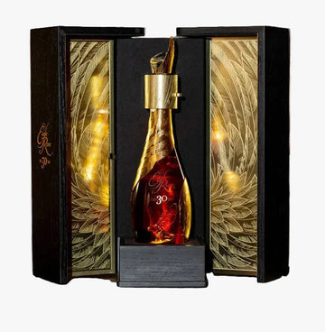 A premium bottle of cognac with a gold decanter cap and red liquid, displayed inside an ornate black and gold gift box on a wooden barrel, with blurred whiskey barrels in the background.