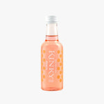 A small clear glass bottle of Kinky Peach Soda Water with pink-orange liquid and orange polka dots, standing on a white surface against a pale peach background.