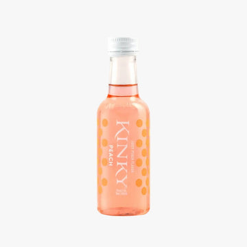 A small clear glass bottle of Kinky Peach Soda Water with pink-orange liquid and orange polka dots, standing on a white surface against a pale peach background.
