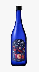 A cobalt blue glass bottle of Moonstone Summer Plum non-alcoholic gin and tonic, standing upright on a rustic wood slice against a textured light gray background.