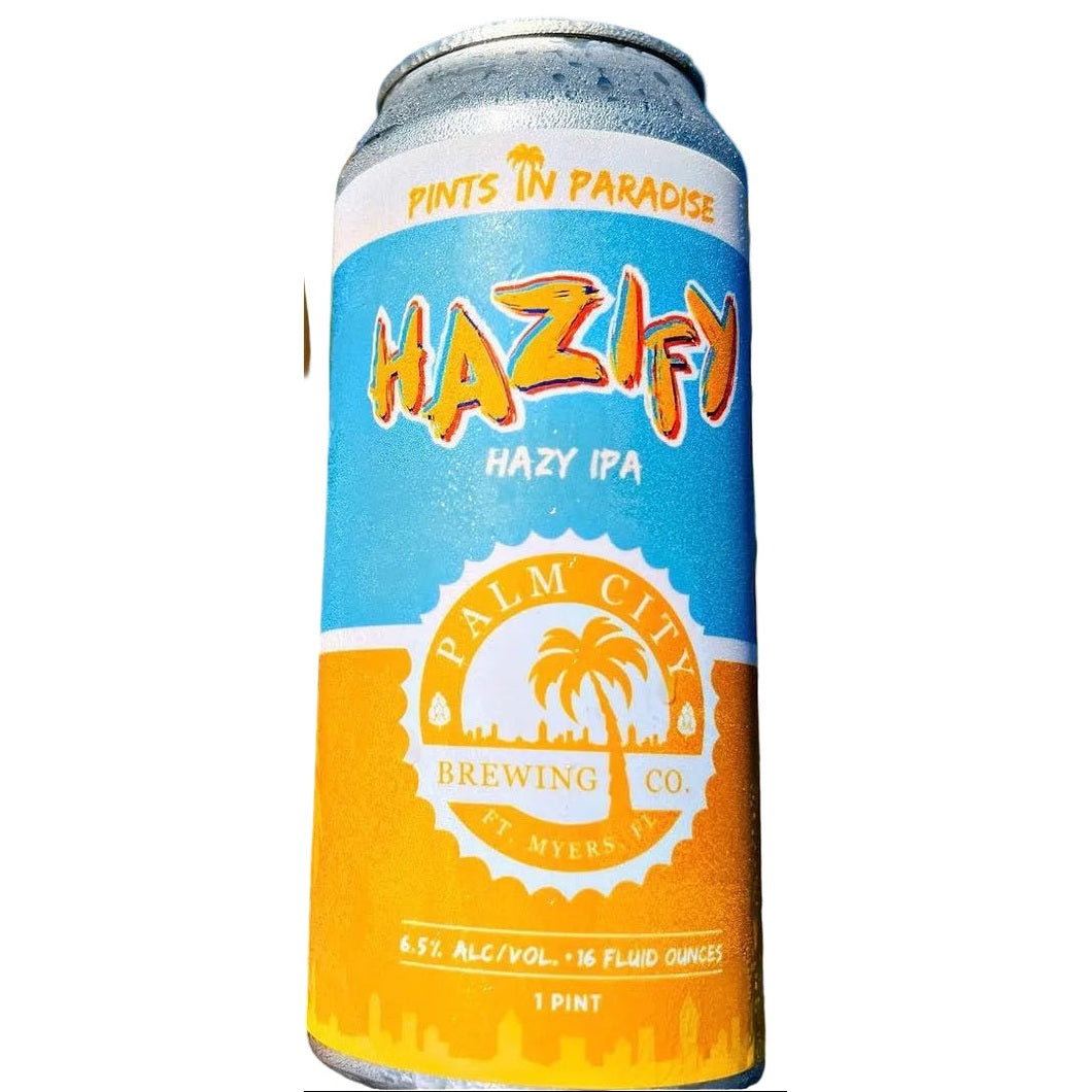 A glass of frothy golden beer and a can of Palm City Brewing Co. Hazify Hazy IPA stand side by side outdoors with palm leaves and blue sky in the background.