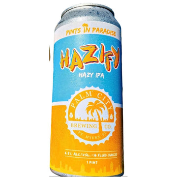 A glass of frothy golden beer and a can of Palm City Brewing Co. Hazify Hazy IPA stand side by side outdoors with palm leaves and blue sky in the background.