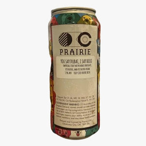 A hand holds a can of Prairie 'You Say Dugal, I Say Hello' sour beer in a brightly lit home interior with a blurred background of framed pictures on a wall.