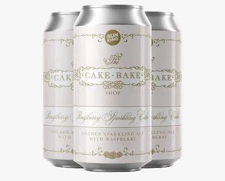 Three silver aluminum beverage cans with white labels and gold decorative text, shown on a pale pink background. The center can reads 'The Cake Bake Shop' and 'Raspberry Sparkling Cider,' with smaller text indicating a collaboration at the top and 'AVAILABLE FEB.