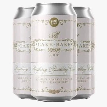 Three silver aluminum beverage cans with white labels and gold decorative text, shown on a pale pink background. The center can reads 'The Cake Bake Shop' and 'Raspberry Sparkling Cider,' with smaller text indicating a collaboration at the top and 'AVAILABLE FEB.