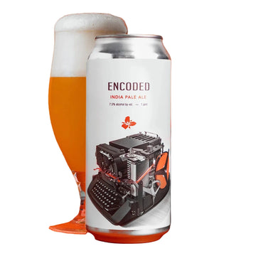 A can of Encoded India Pale Ale stands upright on a bright orange surface, with a filled beer glass behind it and a blurred keyboard prop on the right. The can label is white with dark text and an illustration of a typewriter.