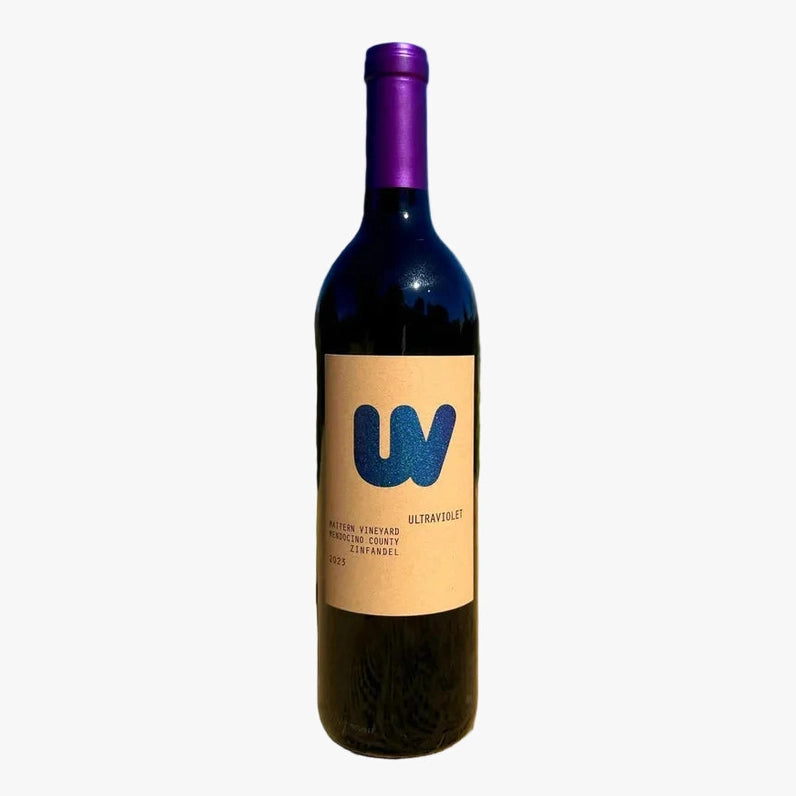 A single bottle of wine standing upright on sandy ground outdoors, with a blurred landscape and blue sky in the background. The bottle has a dark glass body, a purple foil top, and a beige label with a large blue 'W' logo.