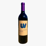 A single bottle of wine standing upright on sandy ground outdoors, with a blurred landscape and blue sky in the background. The bottle has a dark glass body, a purple foil top, and a beige label with a large blue 'W' logo.